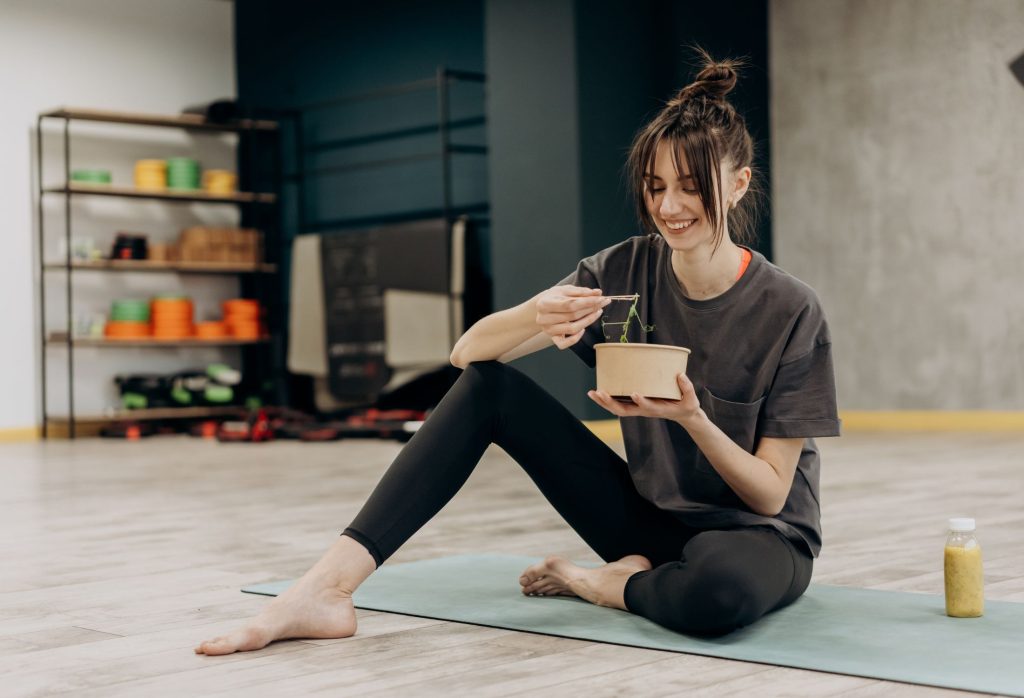 woman sitting on a gym mat eating what looks to be a bowl of noodles or salad.