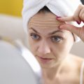 woman examining her forehead wrinkles in a handheld mirror. Her hair is wrapped in a white towel